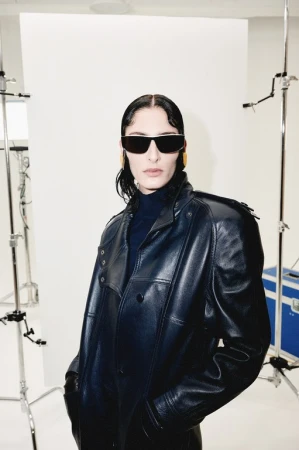 Portrait of a woman with sunglasses and a dark leather coat posing in a studio with white background and light stands.