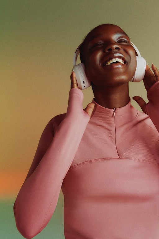 Smiling Black woman with headphones on, wearing a pink fitted long-sleeve shirt in a close up shot with an abstract light background.