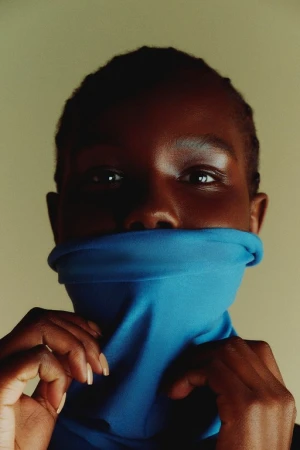 Close-up portrait of a Black woman wearing a bright blue high-neck top pulled up over her lower face, styled by Gemma Hayward.