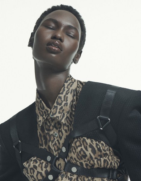 Close-up studio portrait of an adult female model with dark skin wearing an animal print shirt and black blazer with harness straps. 