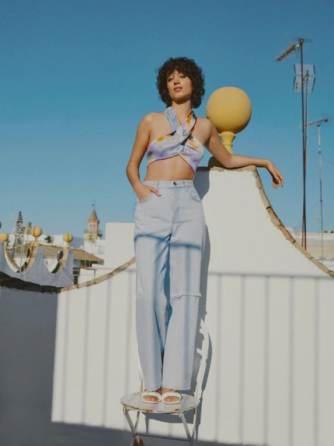 Young woman in halter top and wide-leg jeans strikes a confident pose outdoors on a rooftop, bathed in bright sunlight with blue skies.