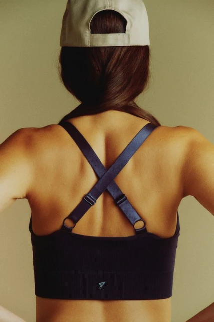 A woman wearing a sports bra and backwards cap with her back to the camera in a studio with a neutral background.