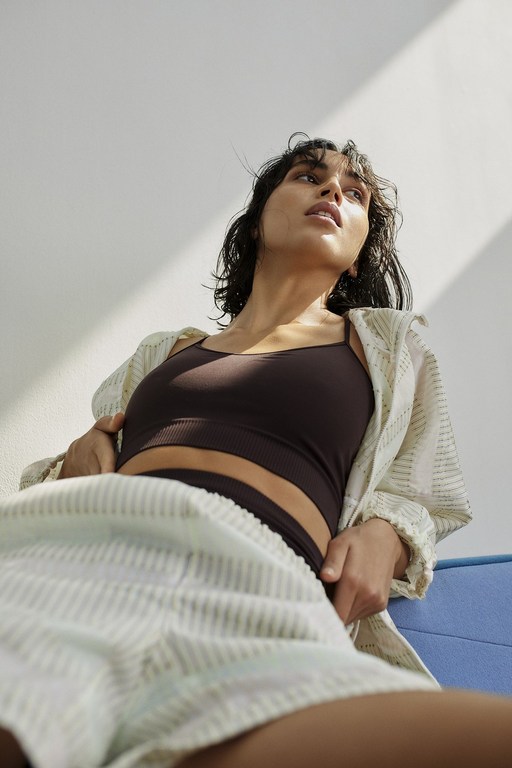 Low angle view of a brunette adult woman in a brown crop top and striped shorts sitting indoors in bright, natural light.