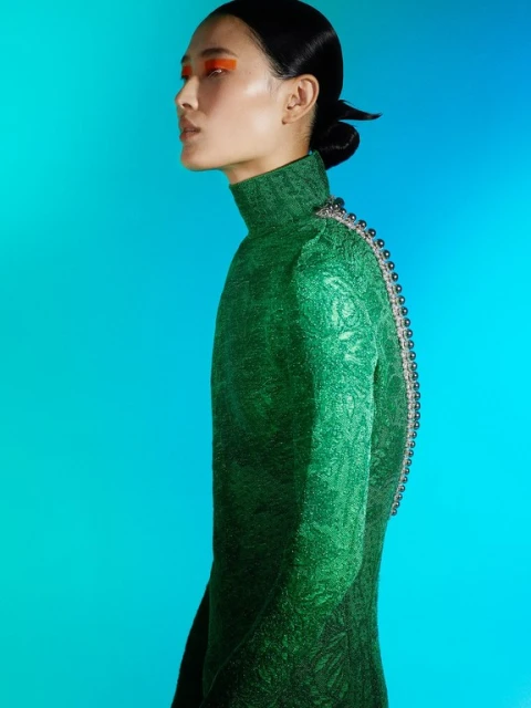 Profile of a woman with slicked back hair in a green textured dress and bright orange eyeshadow against a blue gradient backdrop.