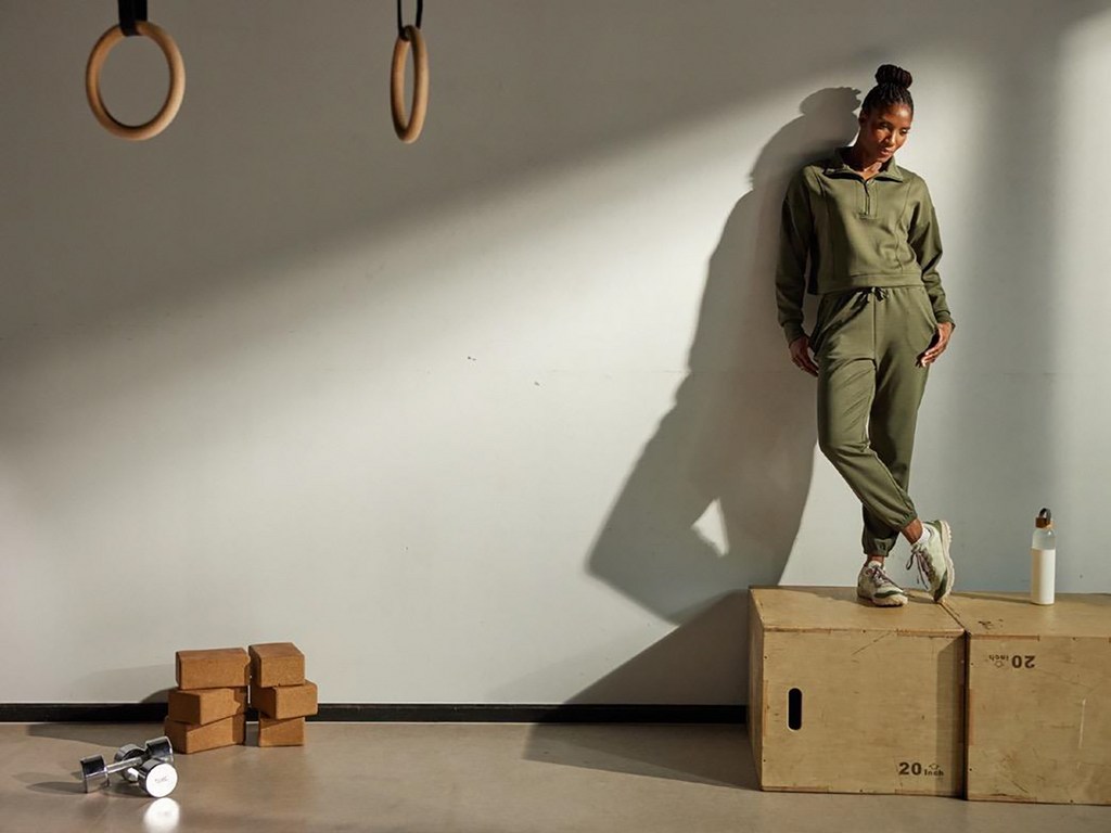 A young Black woman in a matching olive-green sweatsuit stands on a wooden plyometric box in a brightly lit gym setting.