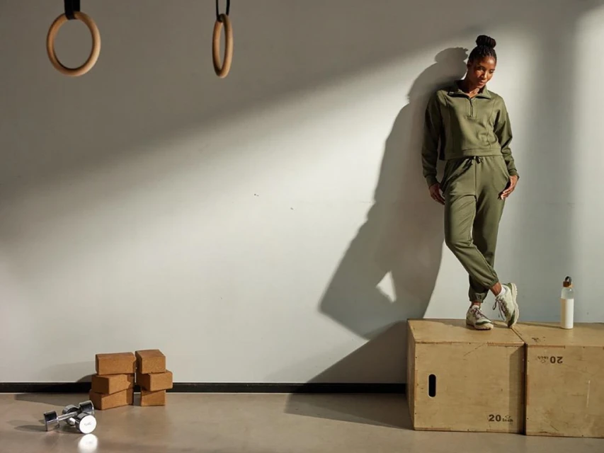 A young Black woman in a matching olive-green sweatsuit stands on a wooden plyometric box in a brightly lit gym setting.