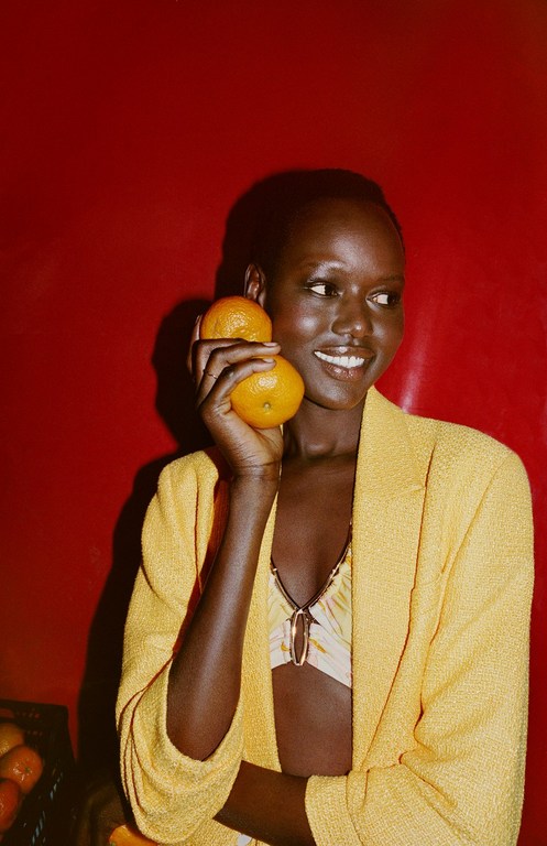 Smiling woman in yellow blazer holds oranges against red backdrop, showcasing a blend of fashion and fresh produce.