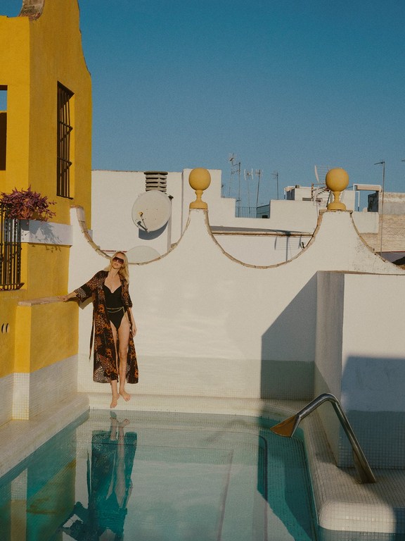 Woman in swimsuit and robe stands on a rooftop next to a pool, framed by white and yellow buildings against a clear blue sky.