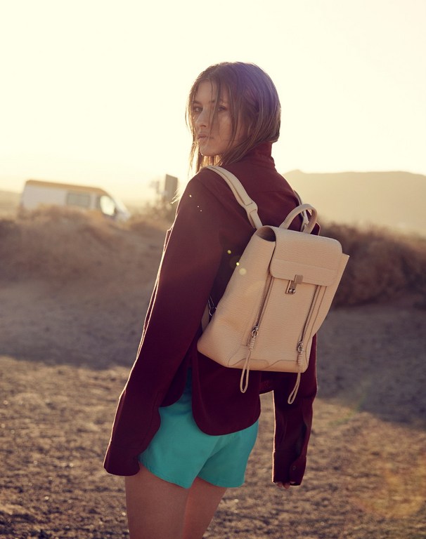 A young woman in a maroon jacket and blue shorts stands outside in a desert landscape, wearing a backpack and looking at the camera.