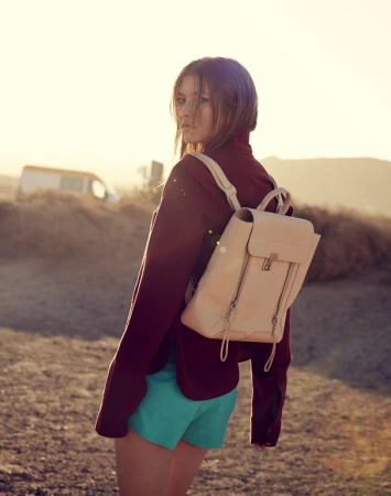 A young woman in a maroon jacket and blue shorts stands outside in a desert landscape, wearing a backpack and looking at the camera.