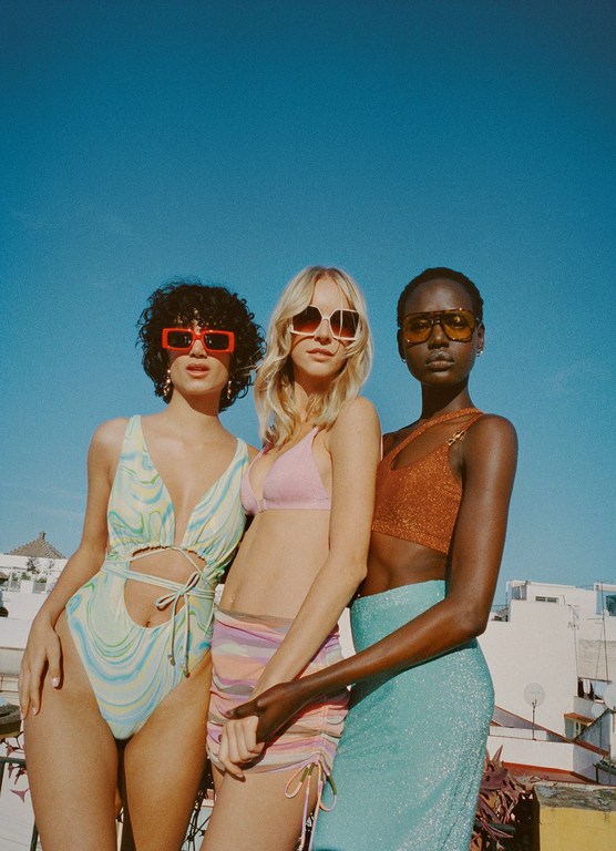 Three fashion models in swimwear and skirts pose outdoors under a bright blue sky, showcasing summer fashion and colorful sunglasses.