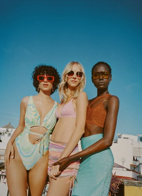 Three fashion models in swimwear and skirts pose outdoors under a bright blue sky, showcasing summer fashion and colorful sunglasses.