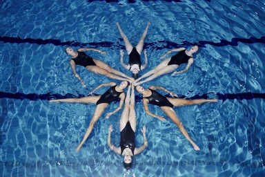 Overhead view of six synchronized swimmers in black swimsuits forming a star shape in a blue swimming pool