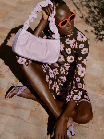 Black model seated outdoors wearing a floral dress, orange sunglasses, and holding a ruched lavender handbag styled by Gemma Hayward.