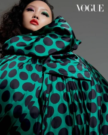 A woman with colorful eye makeup and red lipstick poses in a dramatic green and black polka dot dress with Vogue logo in upper right.
