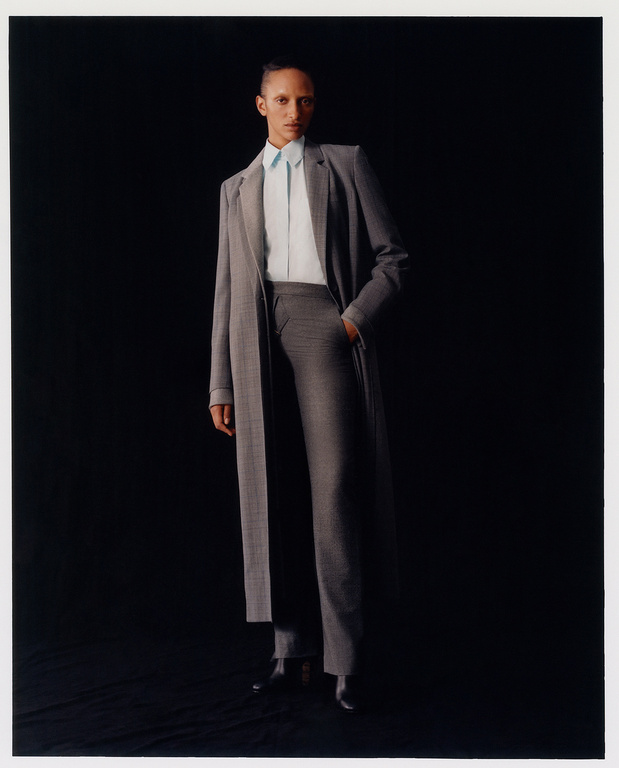 Fashion editorial shot of a single African-descent adult female wearing grey coat and trousers with a blue shirt against black backdrop.