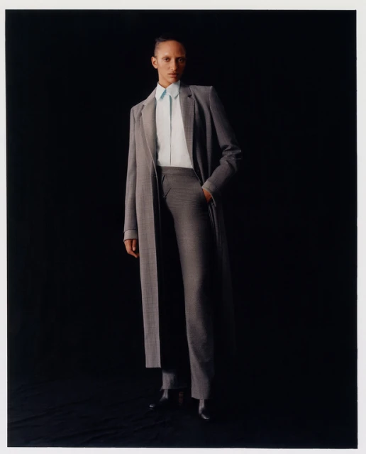 Fashion editorial shot of a single African-descent adult female wearing grey coat and trousers with a blue shirt against black backdrop.