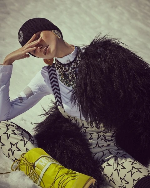 Fashionable woman in stylish winter clothing and accessories poses in snow-covered landscape with mountains in the background on a bright day.