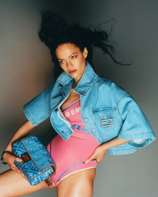 Woman poses in pink bodysuit, denim jacket, and carries a blue handbag, showcasing fashion against a neutral backdrop.