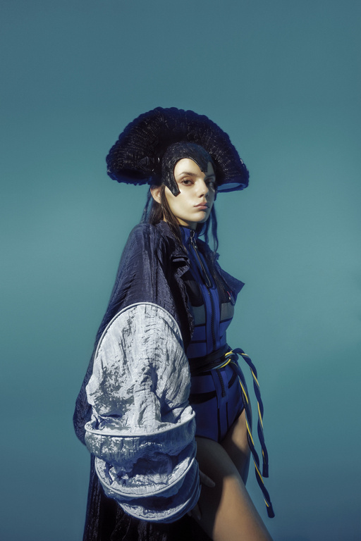 Fashionable young woman in blue futuristic outfit and large black headdress poses in a studio with a light blue background.