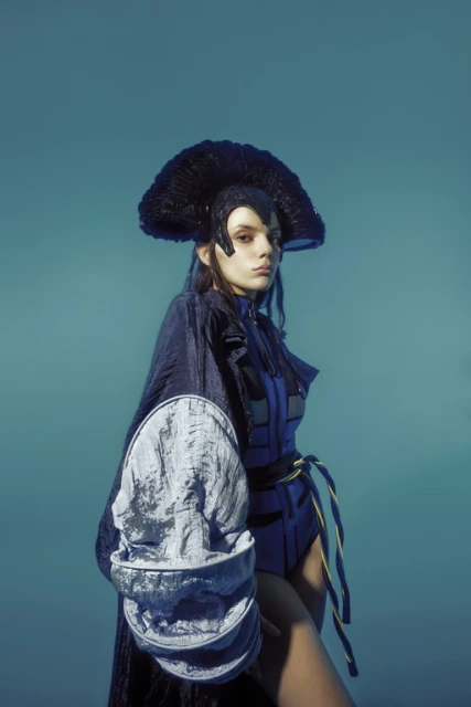 Fashionable young woman in blue futuristic outfit and large black headdress poses in a studio with a light blue background.