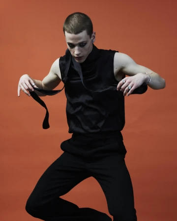 Young man with dark eye makeup wearing a black sleeveless top against an orange background styled by Katie Felstead
