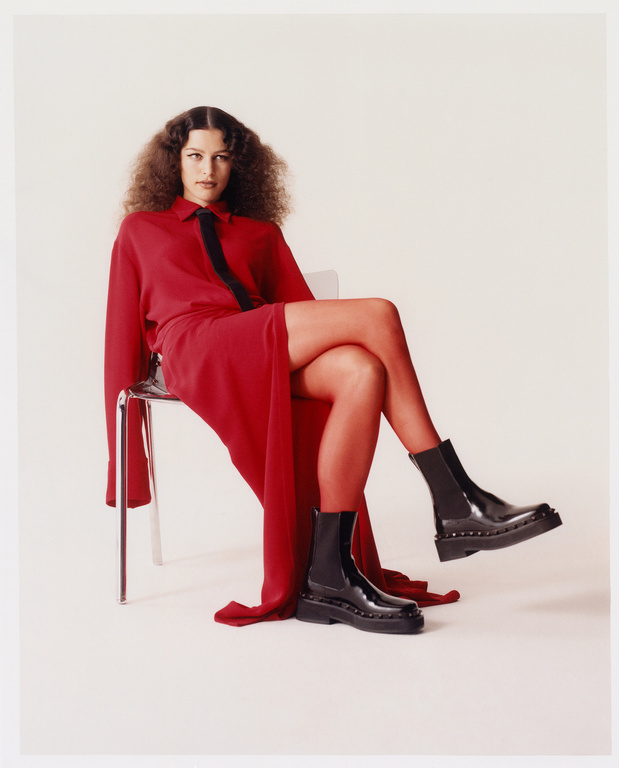 Elegant woman in a red dress and tights sits on a transparent chair wearing shiny black platform boots in a fashion photography studio.