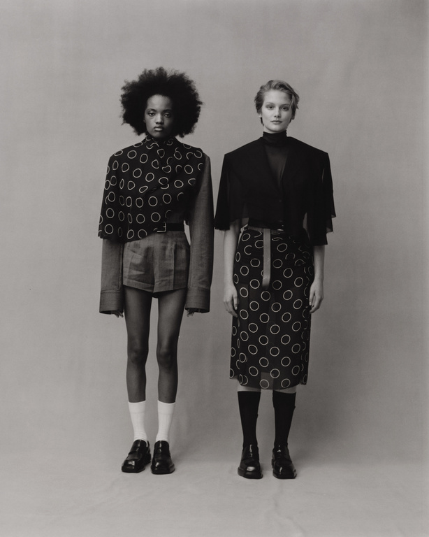 Two fashion models in studio, one Black woman with afro and one White woman with short blonde hair, wearing patterned skirts and jackets.