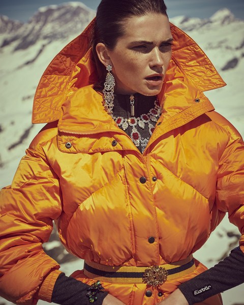 Fashion shot of a woman wearing a bright orange puffer jacket with jewelry posing in front of snow-covered mountains on a sunny day.