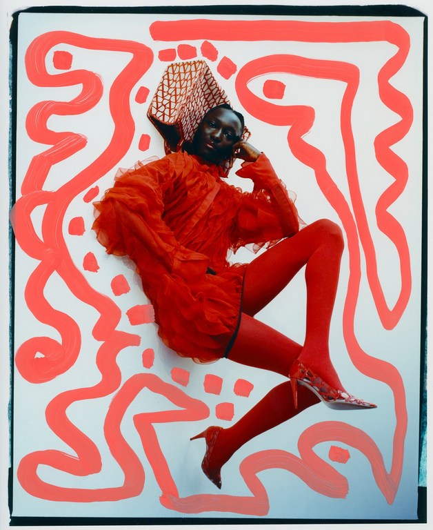 Fashionable black model wearing a red dress, red tights, and heels poses in front of a white background with red paint strokes.