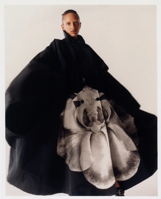 Fashion shot of a Black female model wearing an oversized black coat with a large floral design, posing in front of a plain white backdrop.