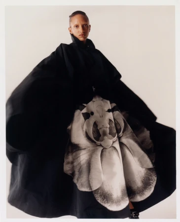 Fashion shot of a Black female model wearing an oversized black coat with a large floral design, posing in front of a plain white backdrop.