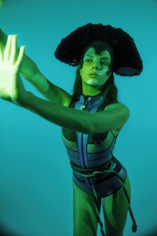 Model in a blue bodysuit and large black headpiece poses in front of a blue background with arms raised, hands angled, and green lighting.