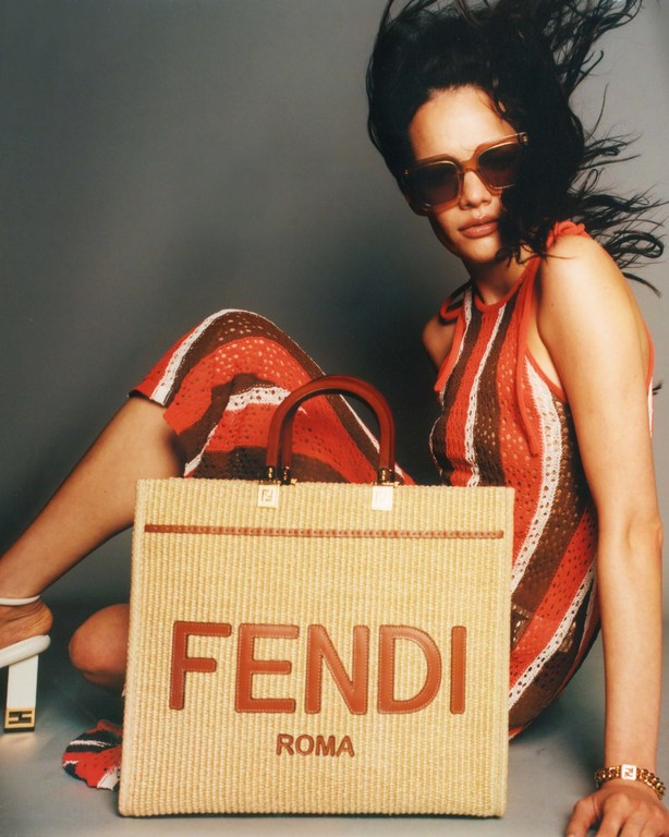 A woman poses indoors with a Fendi straw tote bag featuring leather logo and handles, wearing sunglasses and a striped dress.