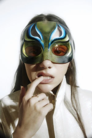 Close-up portrait of a woman wearing an airbrushed patterned mask and a high-collar white jacket, biting her finger.