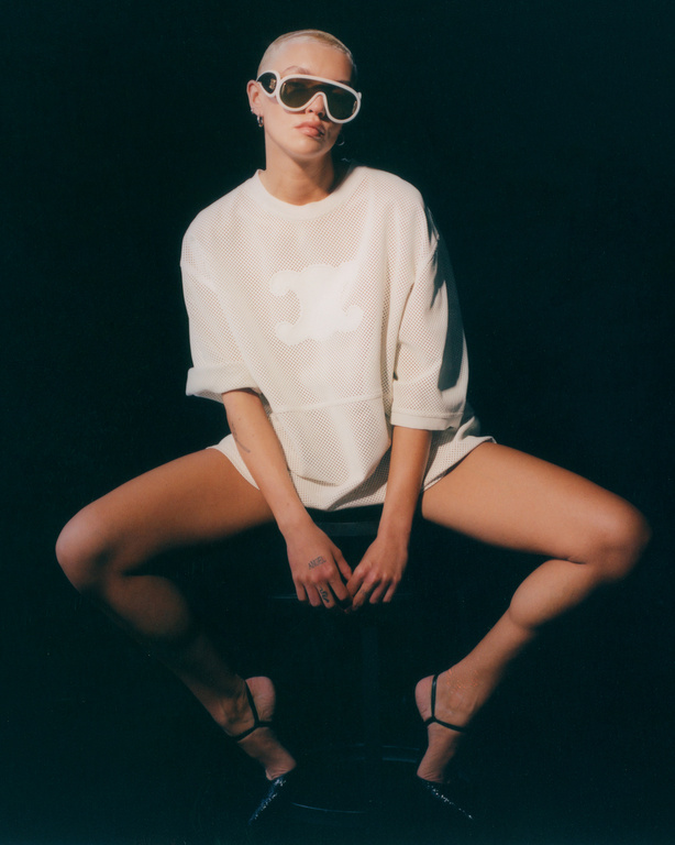A slender adult posing on a black stool wearing white sunglasses, a white mesh shirt and strappy heels against a dark background.