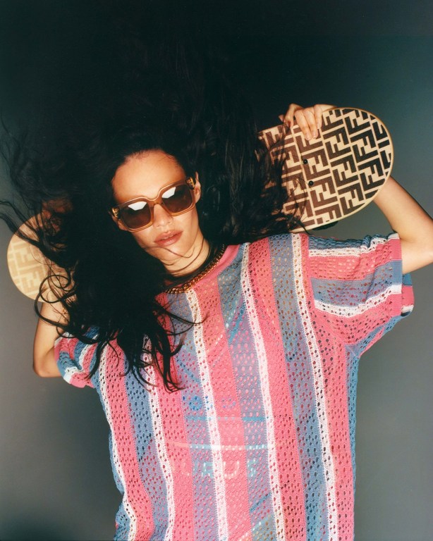 Stylish woman with wavy dark hair wearing sunglasses and a pink and blue striped shirt, holding a patterned handbag overhead.