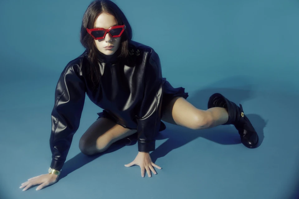 A young woman poses wearing a black dress, black boots and red sunglasses against a blue background in a fashion studio shot.