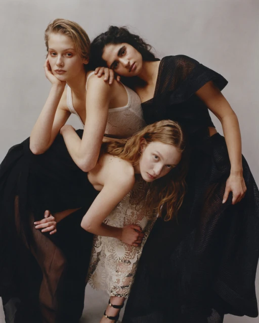Three young women pose in a close-up studio portrait, wearing dresses with mesh and lace details, against a neutral background.