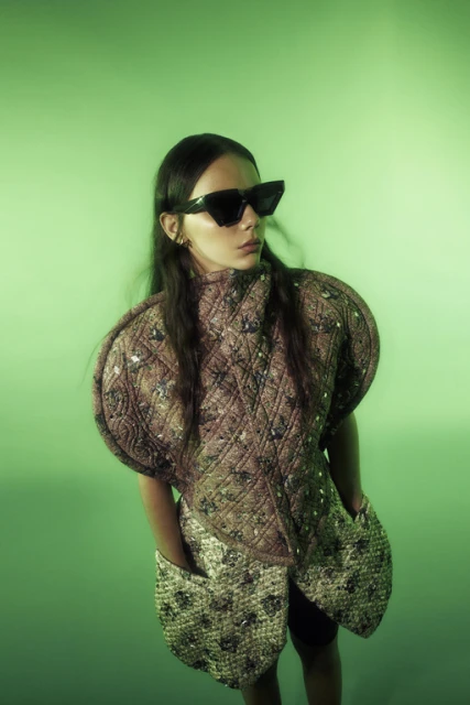Young woman in avant-garde, quilted tan mini dress and dark sunglasses against a green background in a fashion-forward portrait.