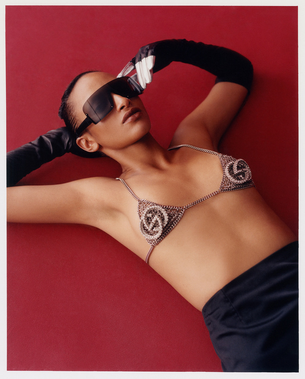 A woman lies against a red backdrop wearing a silver chain bra top with logo detail, black gloves, dark sunglasses, and black pants.