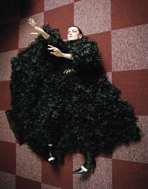 A woman in a large black textured garment reclines on a red-and-white checkered surface with hands raised, creating a fashion editorial.
