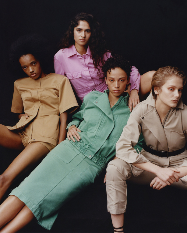 Group of four fashion models in coordinated earth-tone and pastel outfits posing in a studio with a dark background looking at camera.