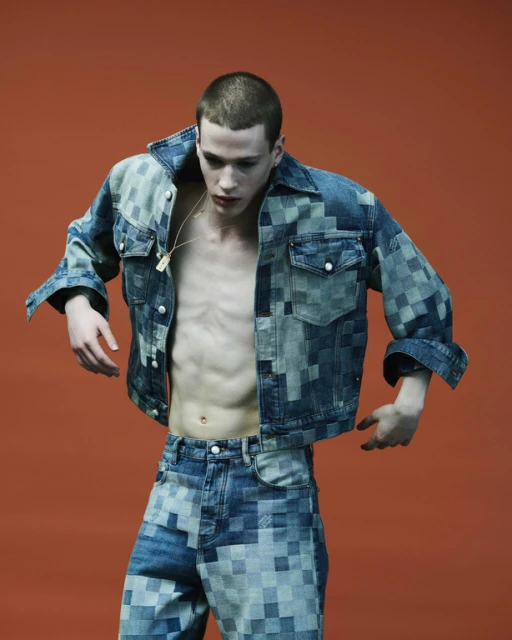 Young man posing shirtless in patterned denim jacket and jeans against an orange studio backdrop, fashion editorial photography.