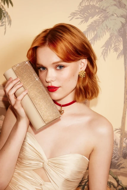 Elegant red-haired woman with clutch. Close-up shot of model with red lips, wearing a cream-colored dress in front of a palm tree backdrop.