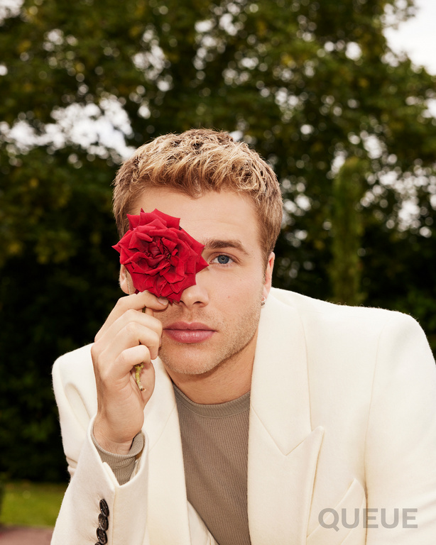 A young adult male in an off-white suit holds a red rose over his eye, standing outdoors with a blurred green background.