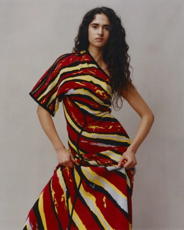 Woman with long dark curly hair wearing a red, yellow, and black patterned dress against a neutral grey studio background