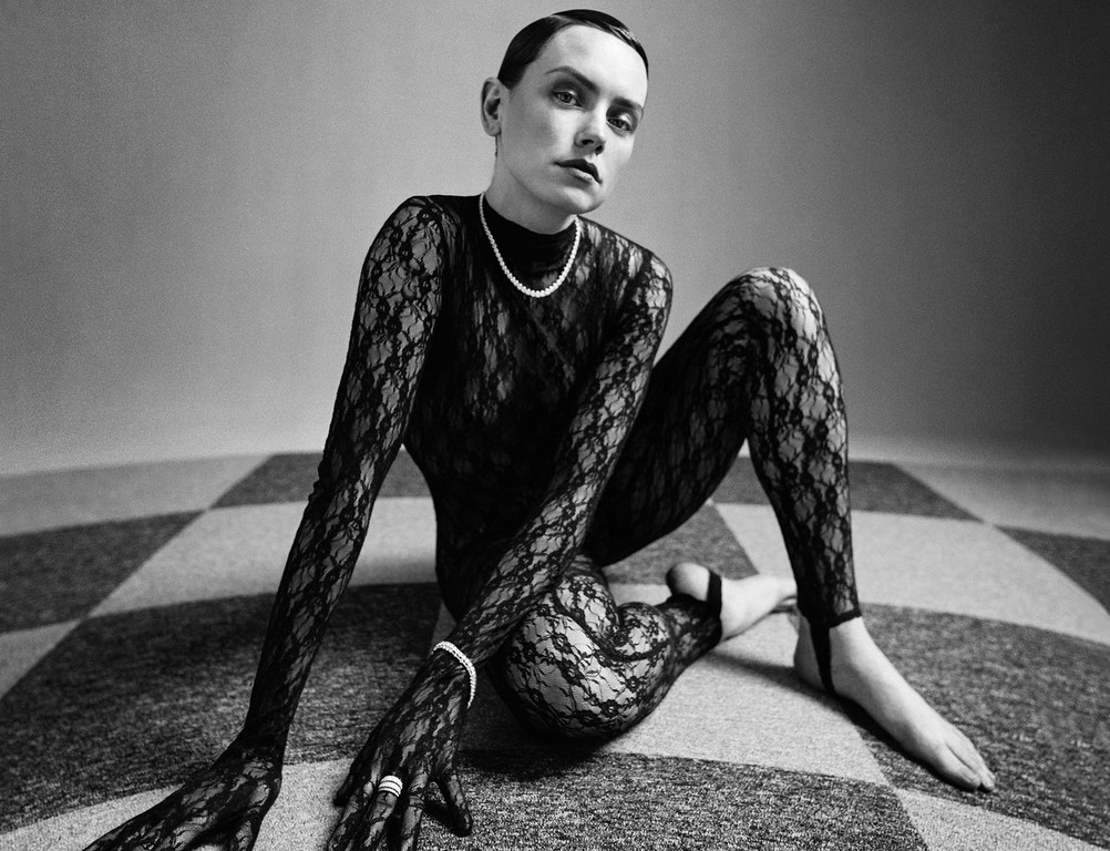 Black and white fashion portrait of a woman in a lace bodysuit and pearl necklace, seated on a checkered surface.