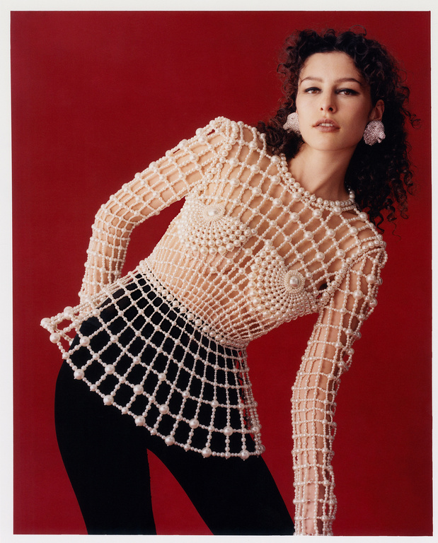 A woman with curly hair poses in a studio wearing a pearl-embellished net top and black pants against a red backdrop in a medium shot.