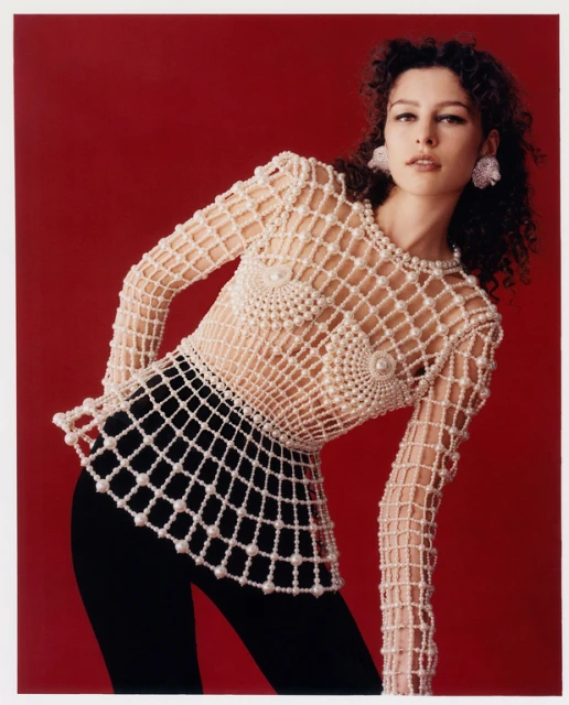 A woman with curly hair poses in a studio wearing a pearl-embellished net top and black pants against a red backdrop in a medium shot.
